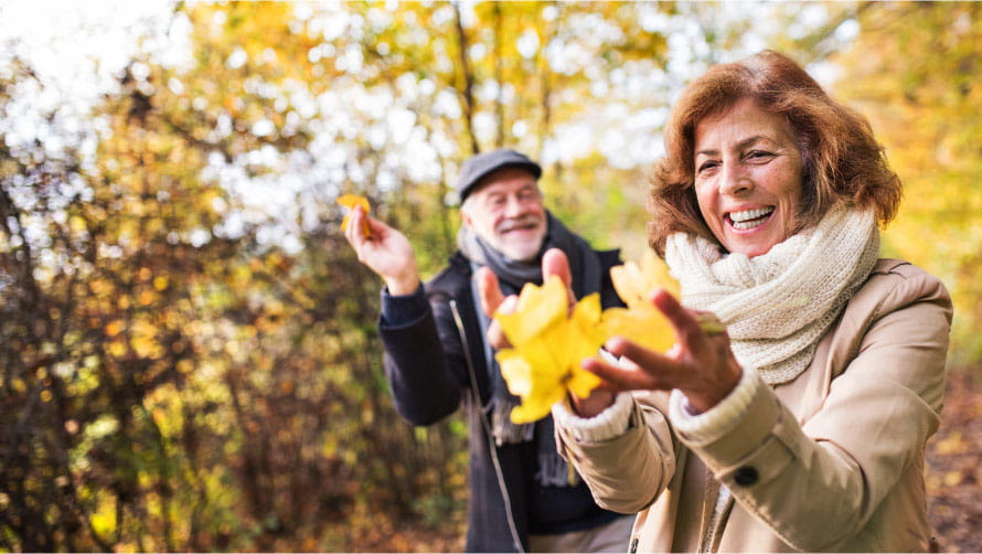 Older couple outside in autumn, the woman is in the foreground with a handful of yellow leaves 