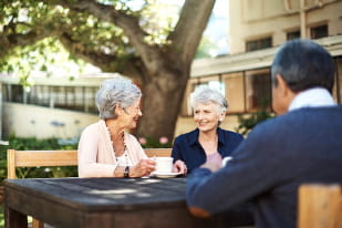 a small group of older friends sitting at a table outside, drinking coffee and chatting