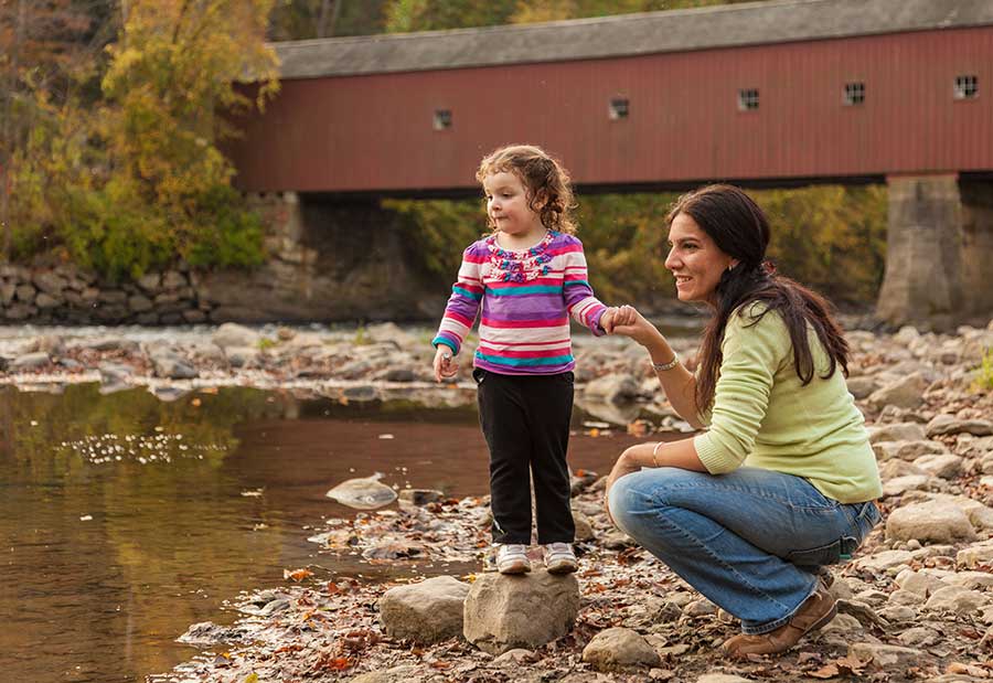 woman with young daughter standing at shore of creek with covered bridge in the background