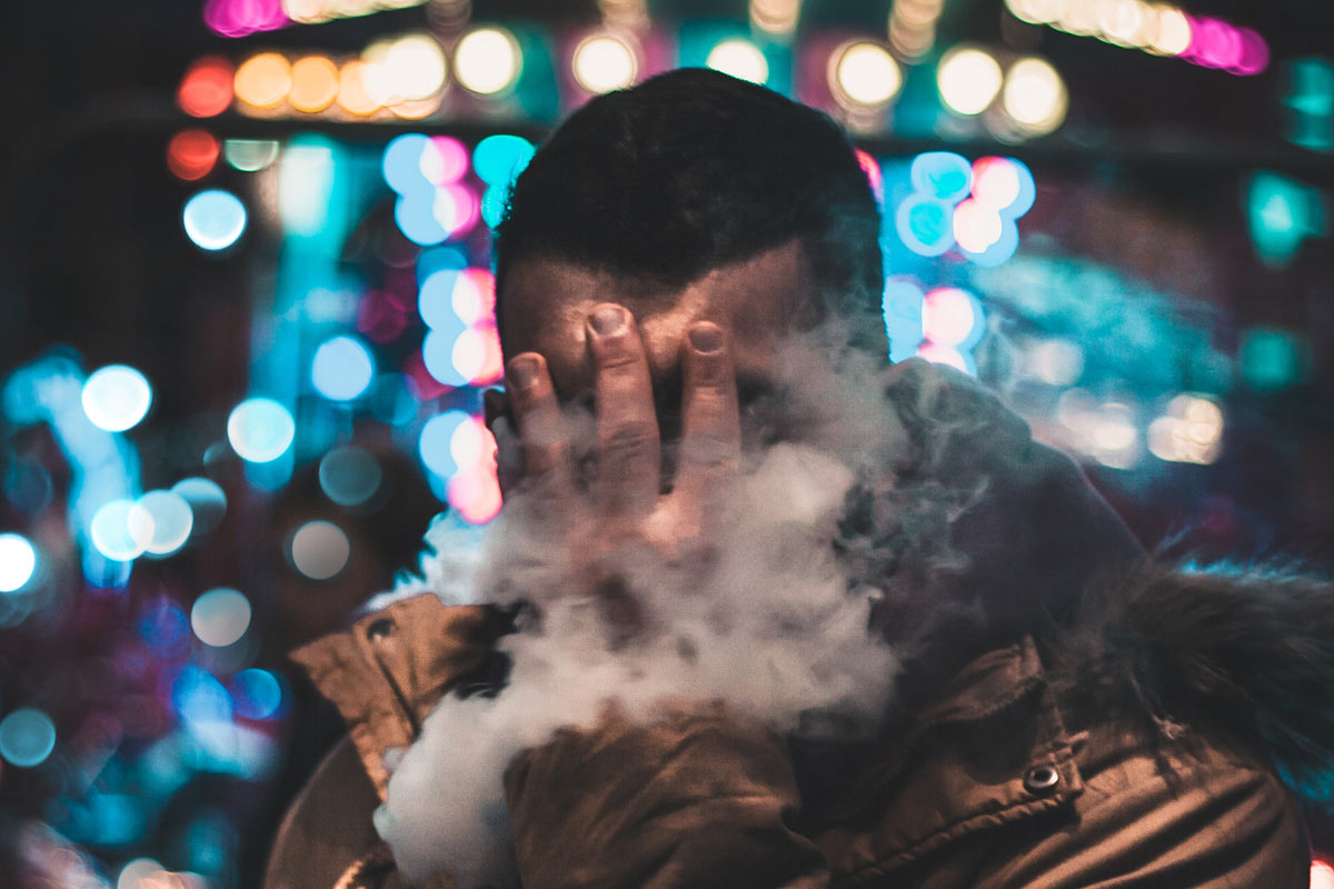 person with hand covering their face and exhaling vapor from pen