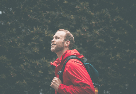 Man in red jacket with backpack standing against foliage wall