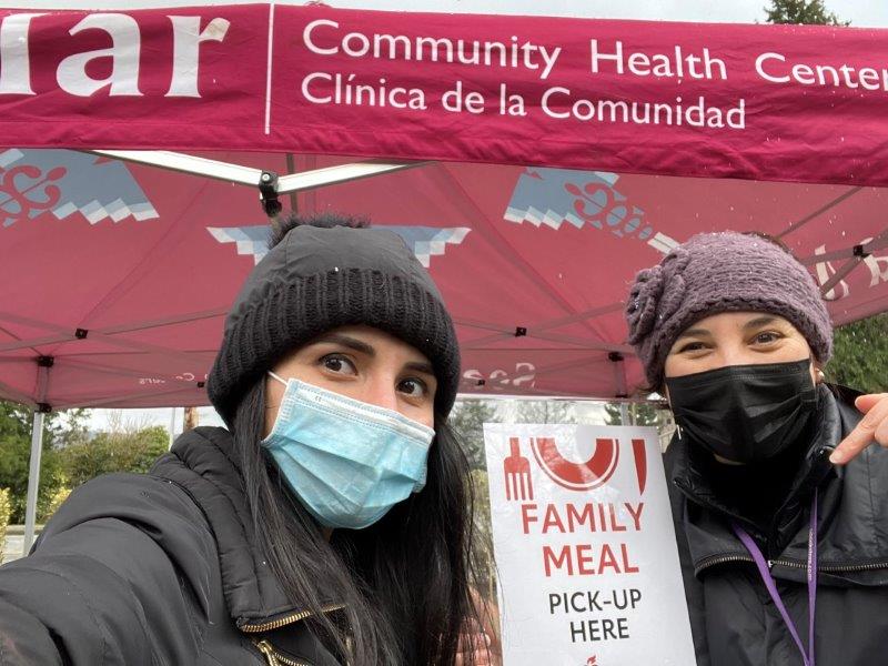 two women wearing masks pose in front of sign: Family Meal Pick-up Here