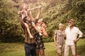 A young couple plays with their child in a grassy park while the grandparents watch nearby