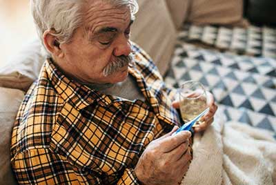 sick elderly man sitting on couch while looking at thermometer