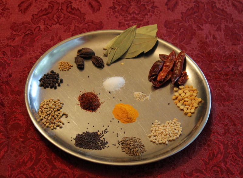 Common spices and condiments used by Saroja Voruganti in her cooking and, in general, in Indian cooking. Clockwise from the top: bay leaf, red chiles, split chickpeas, black gram, cumin seeds, mustard seeds, coriander seeds, black pepper, fenugreek seeds and black cardamom. The inner circle contains salt, asafetida powder, turmeric powder and red chili powder. (Photo courtesy of Saroja Voruganti)