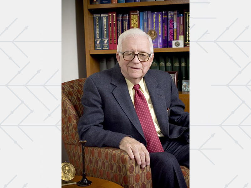 Eugene Braunwald seated beside a bookshelf filled with books.