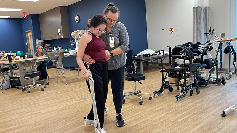 Bella Chambasis receiving support from physical therapist Jadean Hoff as she works on walking with a cane