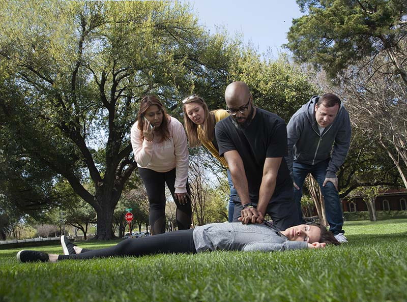 A bystander performs CPR on a woman lying on the grass in a park.