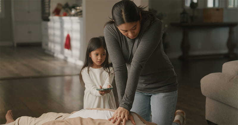 a woman performing CPR on a family member in the living room at home while her young daughter helps by holding her cell phone