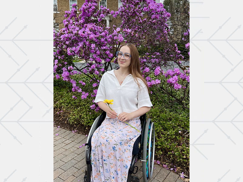 Caroline Laubach smiling and holding a flower, sitting in her wheelchair in front of purple flowering bushes