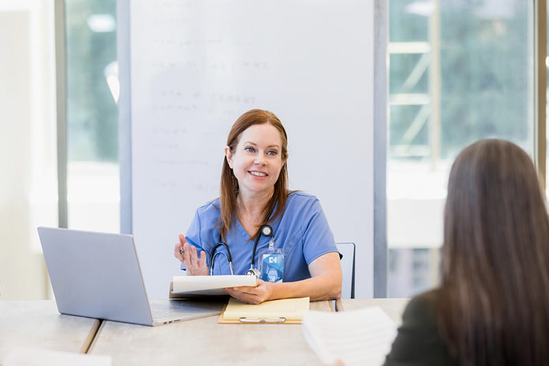 Healthcare professional smiles at colleague while sitting in front of laptop with a clipboard