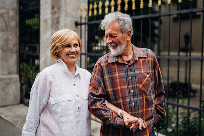 Older couple walking arm in arm along iron fence outside, laughing and smiling
