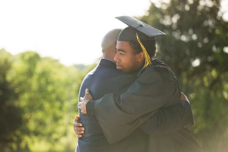male graduate in cap and gown hugging person