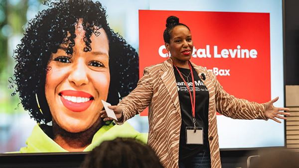 Crystal Levine with CodaBax is speaking to an audience in front of a large digital display. Crystal Levine with CodaBax is speaking to an audience in front of a large digital display.