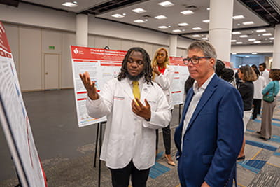 An American Heart Association Undergraduate Scholar is presenting his poster to a Symposium attendee.