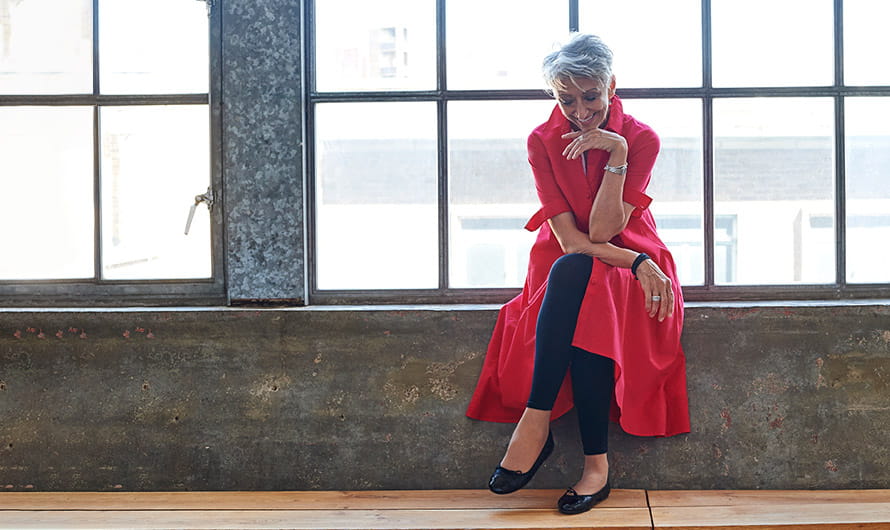 Chic older woman wearing a long red jacket is sitting in front of a large loft window.
