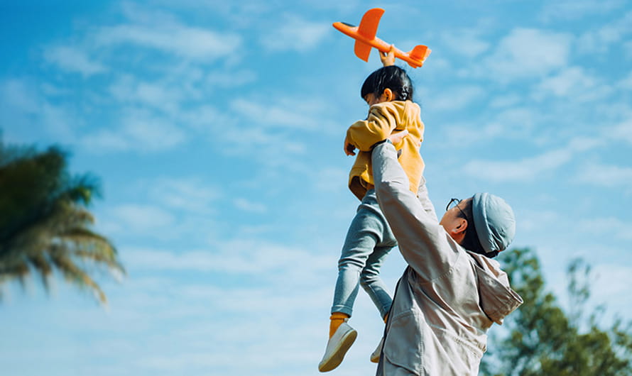 Father lifting daughter up in the air while playing with a toy airplane outside against blue sky