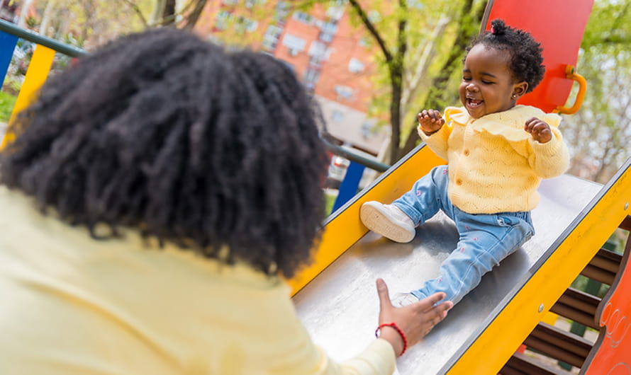 A mother has her hands out to catch her baby daughter on a slide in an outdoor playground.