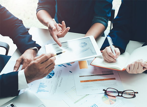 Close up on three people's hands gesturing toward a tablet over a desk with various documents spread out
