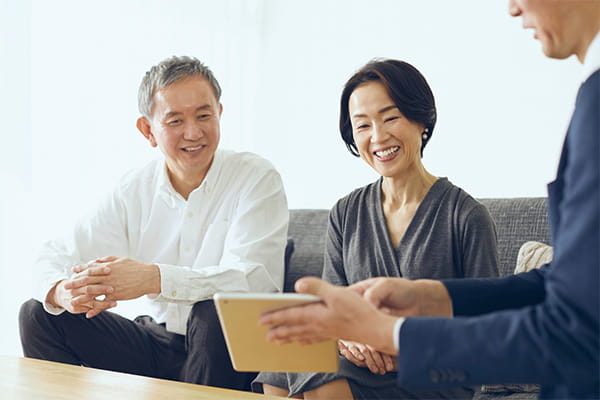 an older couple sitting on a couch and smiling as they look at a tablet being shown to them by an advisor