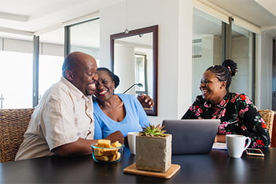 A smiling older couple sitting at a kitchen table with a professional advisor, talking together and looking at a laptop