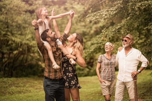 an older couple watches as a young adult couple plays with their child on their shoulders