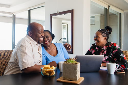 A smiling older couple sitting at a kitchen table with a professional advisor, talking together and looking at a laptop