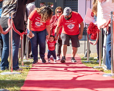 Two parents leaning over to help their small child walk down the Heart Walk red carpet at an event