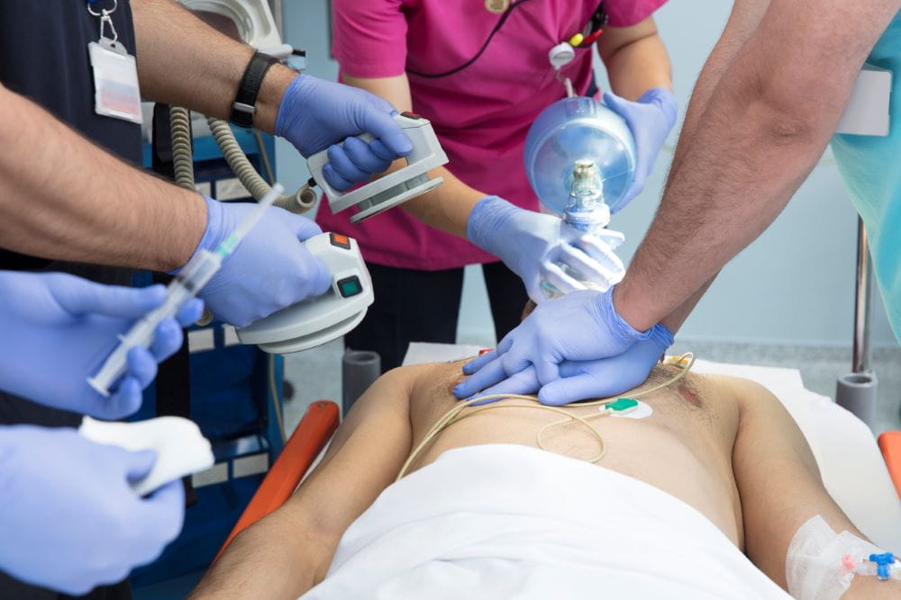 Image of 4 sets of medical workers hands performing airway and paddle resuscitation on a male patient