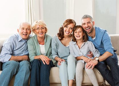 a family of five--two grandparents, two parents, and a young daughter--sit together on a couch, smiling and posing for a photo