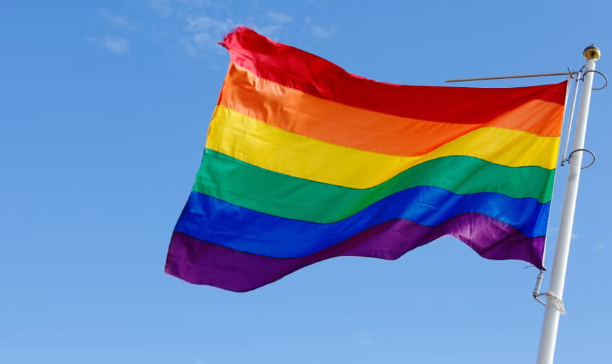 A rainbow flag flying on a flagpole against a blue sky. A rainbow flag flying on a flagpole against a blue sky.